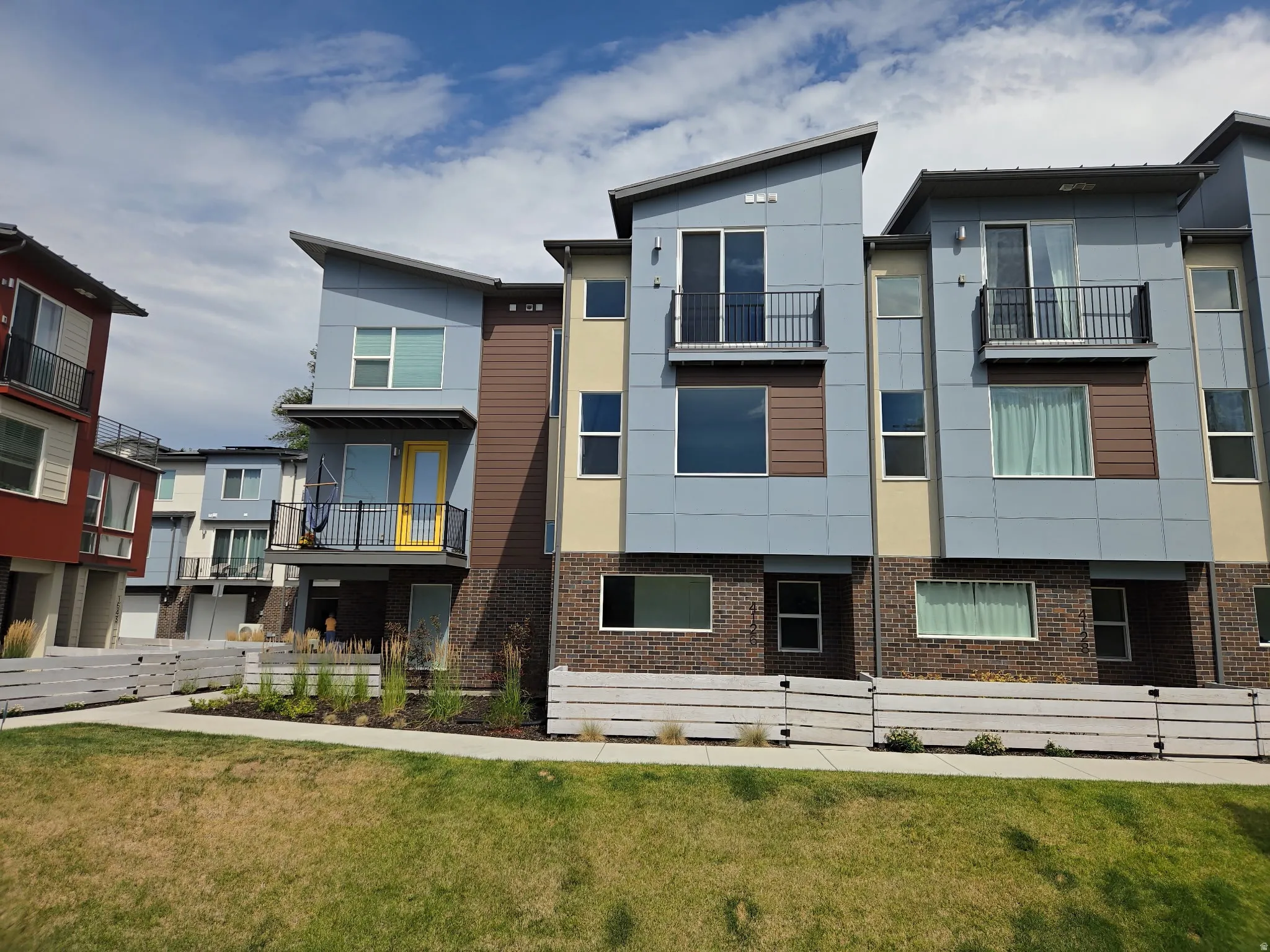 Modern multi-story facade featuring varied siding panels, prominent balconies with black railings, a bright yellow entry door, and a ground-level brick base