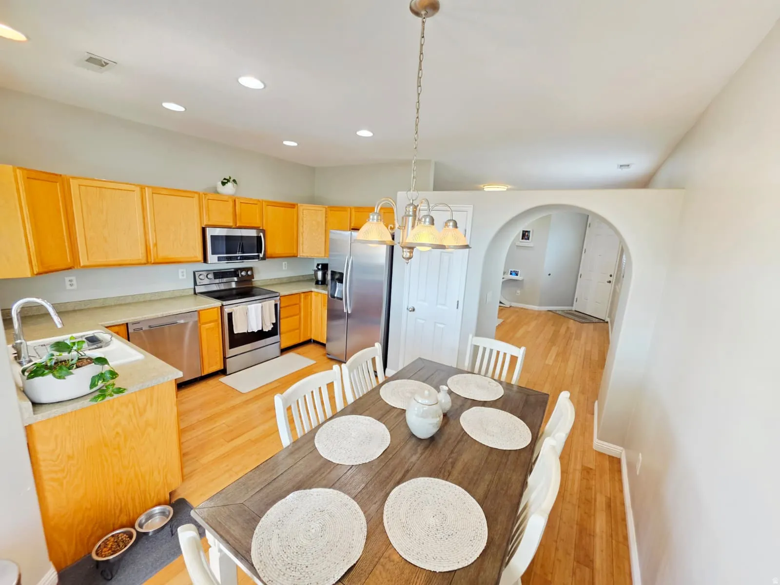 Kitchen and dining area featuring wood-finish flooring, light wood cabinetry, stainless steel appliances, a double basin sink, and an arched doorway