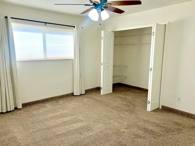 Spacious room featuring neutral carpeting, a dual-door closet with wire shelving, a window with roller blinds, and a ceiling fan with integrated lighting