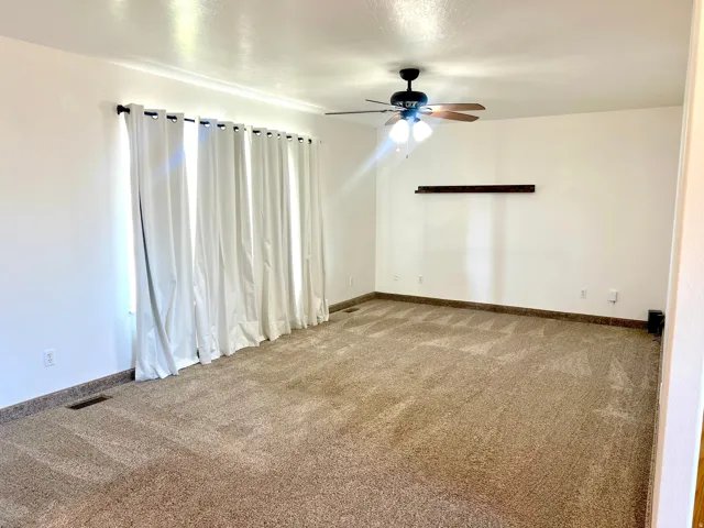 Spacious carpeted room featuring a ceiling fan, a window with white curtains, white walls, and a built-in wall shelf