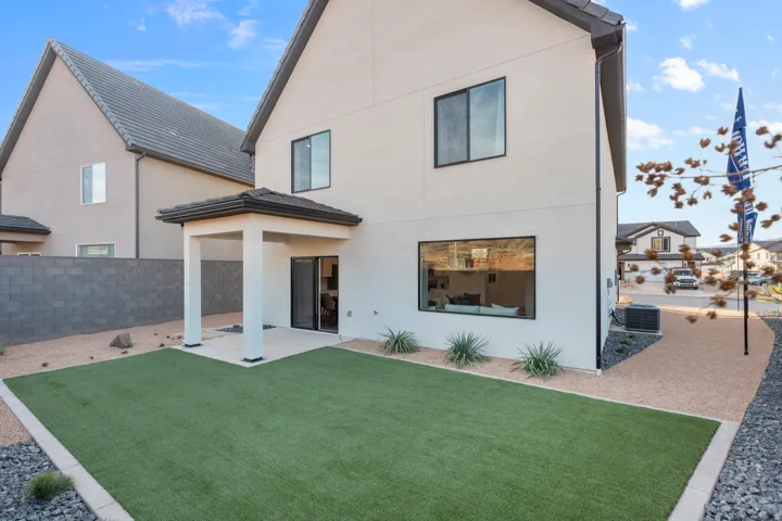 Rear exterior showcasing a covered patio, artificial turf lawn, xeriscaping, and a large horizontal window