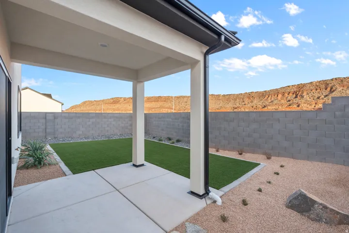Covered patio with integrated downspout, leading to a landscaped backyard featuring artificial turf, decorative gravel, and a block wall perimeter