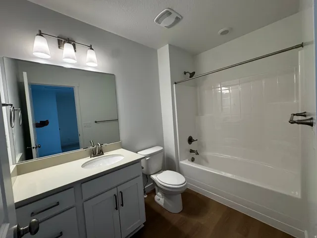 Contemporary bathroom featuring a single vanity with a light-toned countertop, a built-in tub/shower combination, dark bronze fixtures, a wall-mounted light fixture, and wood-finish flooring