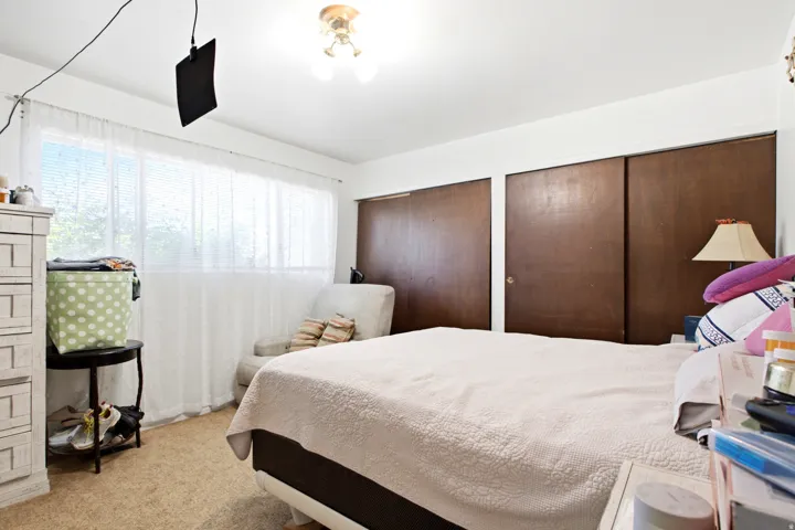 Bedroom featuring light-colored carpet, a wide window with white blinds and sheer curtains, two dark wood-finish sliding closet doors, and a ceiling-mounted light fixture