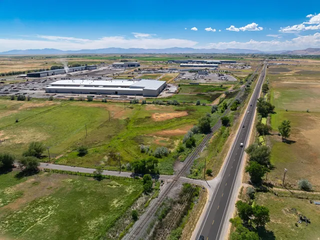 Expansive land parcel featuring a paved road, railroad tracks, and mature trees, surrounded by open fields and distant mountains