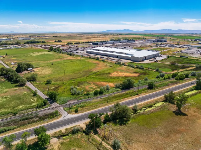 Expansive rural landscape featuring a paved road, railroad tracks, and distant industrial buildings under a clear blue sky