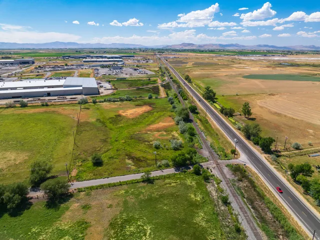 Expansive agricultural fields with distant mountains under a blue sky, featuring a paved road, railroad tracks, and industrial buildings in the background