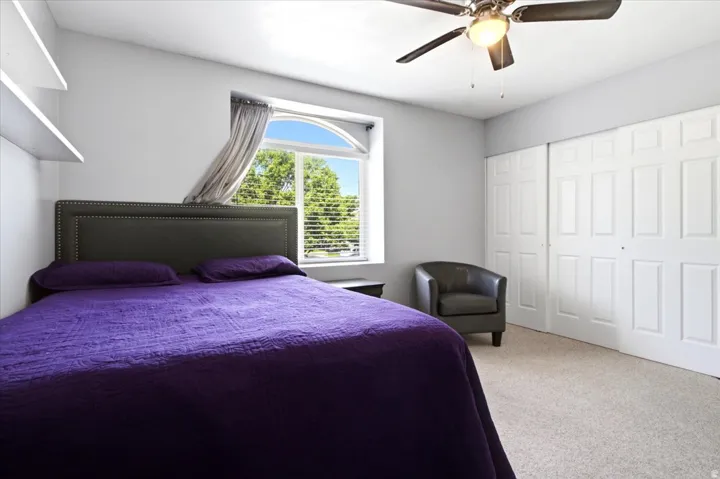 Bedroom featuring a large arched window, a ceiling fan with integrated lighting, two white sliding closet doors, built-in shelving, and neutral wall-to-wall carpeting