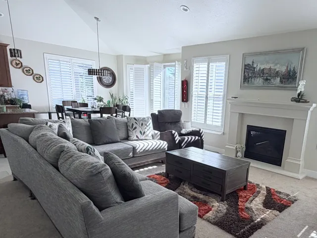Spacious living area featuring neutral carpeting, a built-in fireplace with a stone surround and mantel, and multiple windows with plantation shutters
