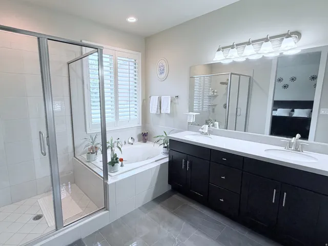 Bathroom featuring a dual vanity with dark wood cabinetry, a built-in soaking tub, a glass-enclosed shower, and plantation shutters