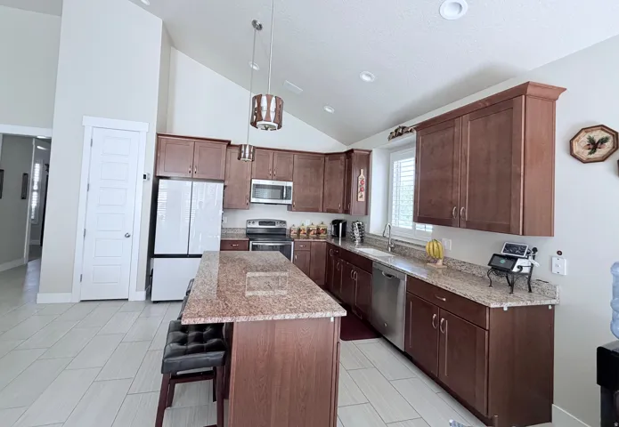 Kitchen featuring vaulted ceilings, wood-finish cabinetry, stainless steel appliances, granite countertops, and light-toned tile flooring