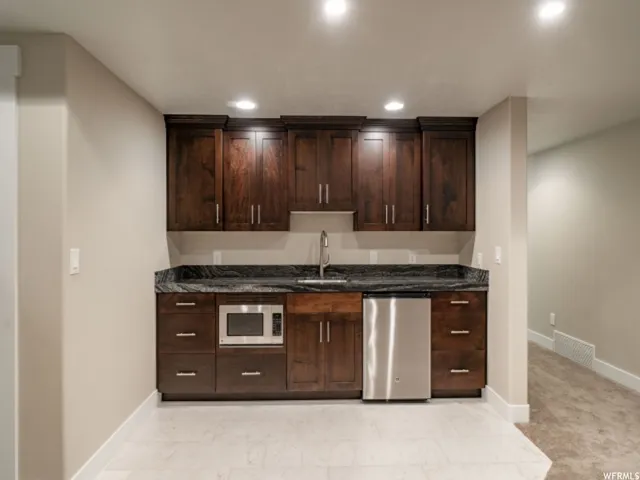 Kitchenette featuring dark wood cabinetry, a built-in microwave, stainless steel dishwasher, and dark stone countertops