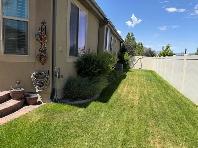 Expansive side yard featuring a vinyl privacy fence, lush green lawn, mature perimeter plantings, and a brick-finish patio area