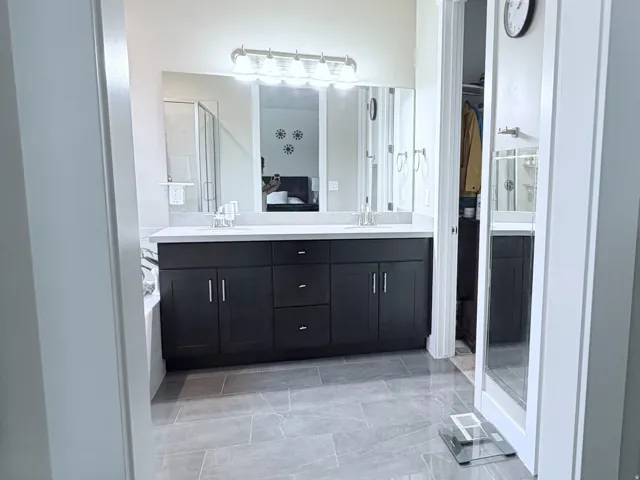 Bathroom featuring expansive tile flooring, a dual vanity with dark cabinetry and a light-toned countertop, a large mirror with overhead lighting, and a glass-enclosed shower