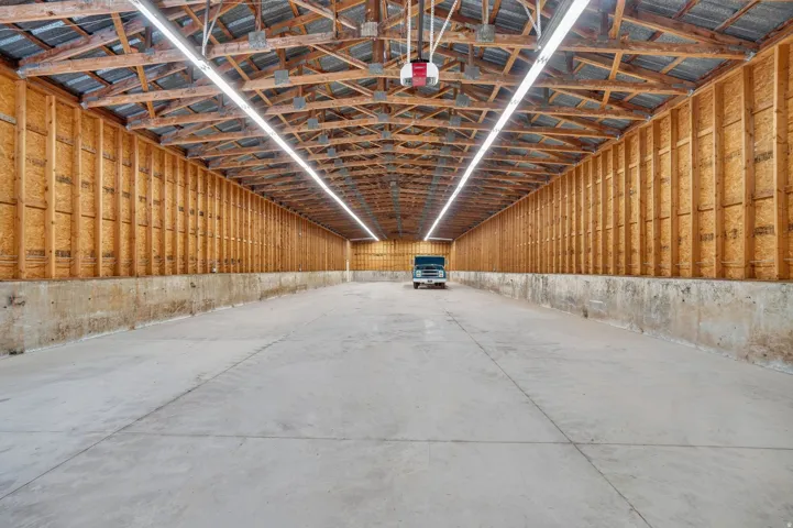Spacious structure featuring a concrete floor, exposed wooden ceiling trusses, and bright overhead strip lighting