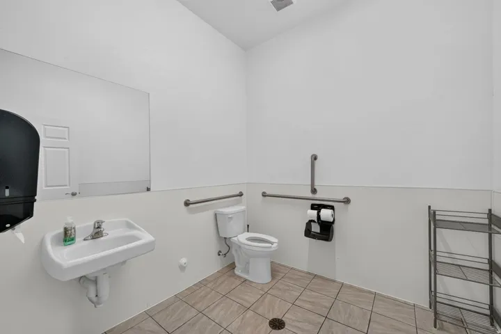 Wall-mounted sink with stainless steel faucet, white toilet, grab bars, and beige tiled flooring