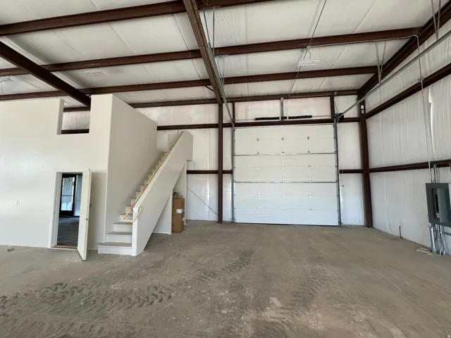 Spacious open warehouse area featuring a large overhead door, exposed structural steel beams, a high insulated ceiling, concrete flooring, and an interior staircase leading to an upper level