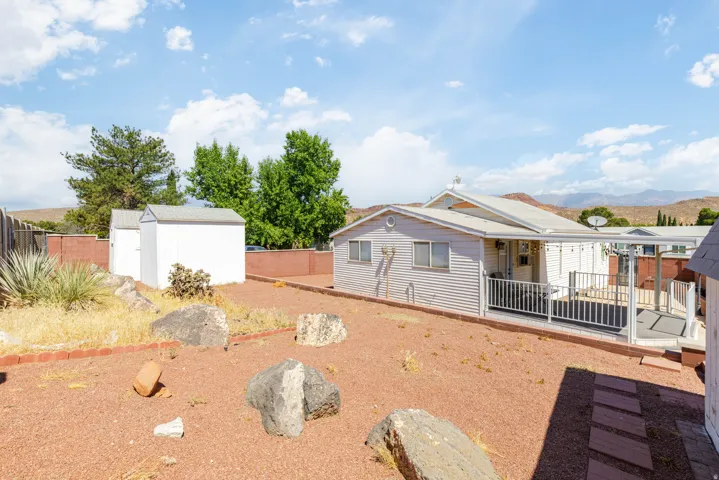 Spacious backyard featuring a covered patio with white railing, red brick retaining wall, and two white storage sheds