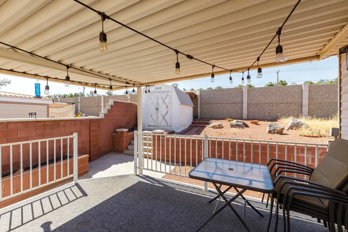 Covered patio featuring string lighting, white railings, a block wall planter, and access to a terraced, low-maintenance yard with a storage shed