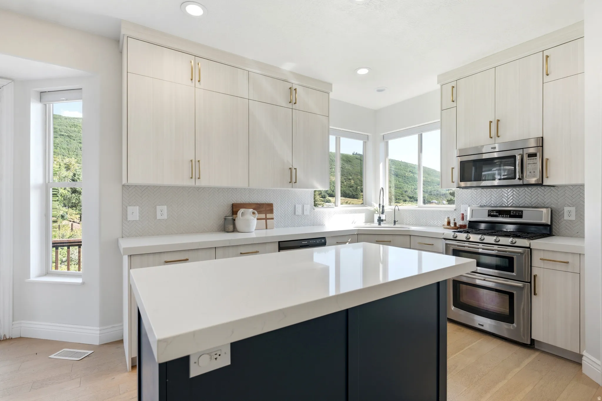 Modern kitchen featuring a central island with a light-toned countertop, light wood-finish cabinetry, stainless steel appliances, and a herringbone pattern tile backsplash