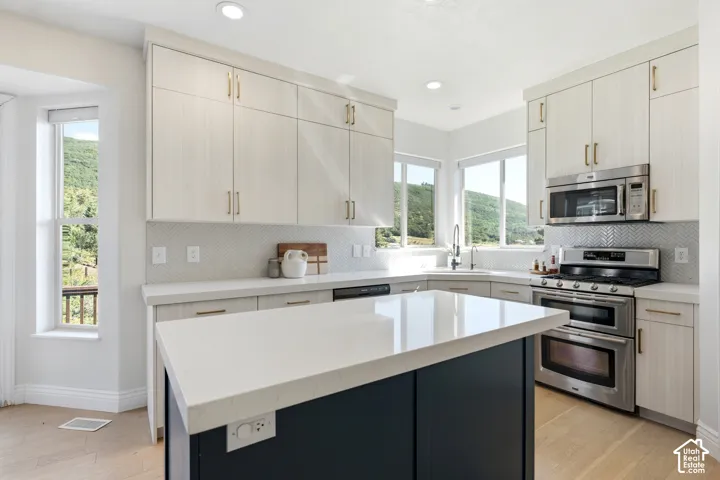 Modern kitchen featuring a central island with a light-toned countertop, light wood-finish cabinetry, stainless steel appliances, and a herringbone pattern tile backsplash