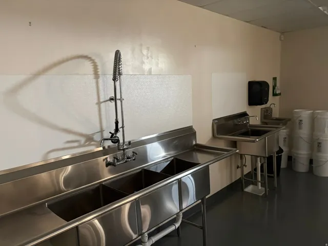 Commercial kitchen space featuring a large stainless steel triple-basin sink with a pre-rinse faucet, a single-basin handwashing sink, and a separate double-basin utility sink