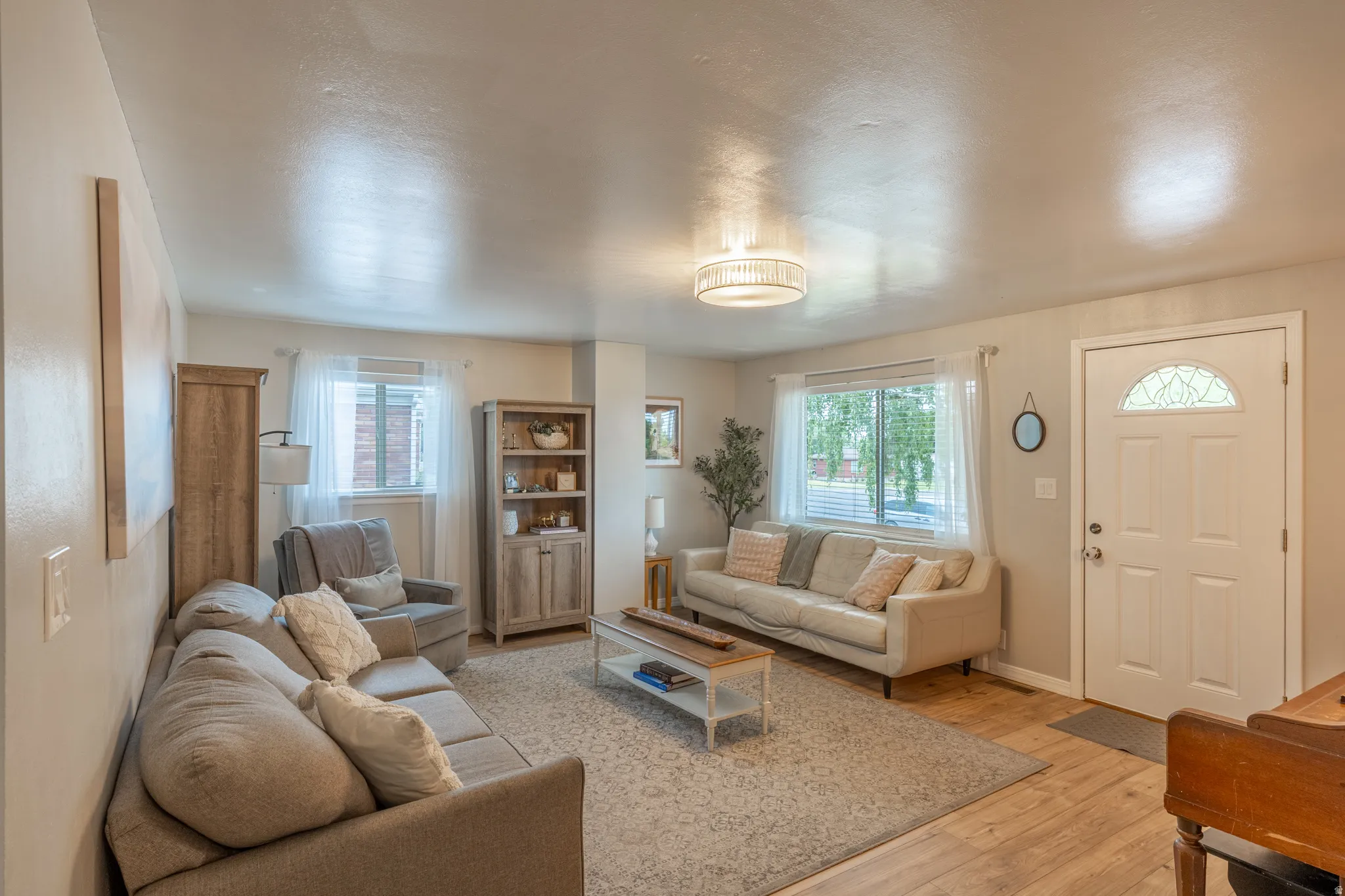 Spacious living area featuring light wood-finish flooring, a decorative ceiling fixture, and multiple windows with white trim