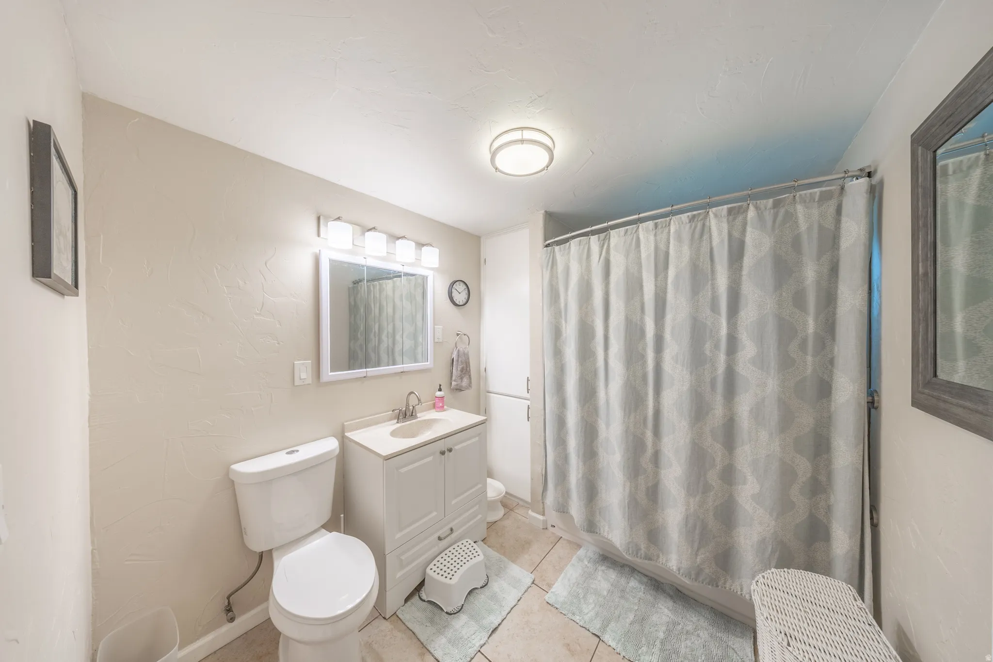 Bathroom featuring a white vanity with a light-colored countertop, an overhead mirror with a four-bulb fixture, and a toilet with a visible water supply line