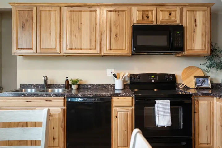West unit. Kitchen featuring light wood cabinetry, dark speckled countertops, and a black appliance suite including a built-in microwave, range, and dishwasher