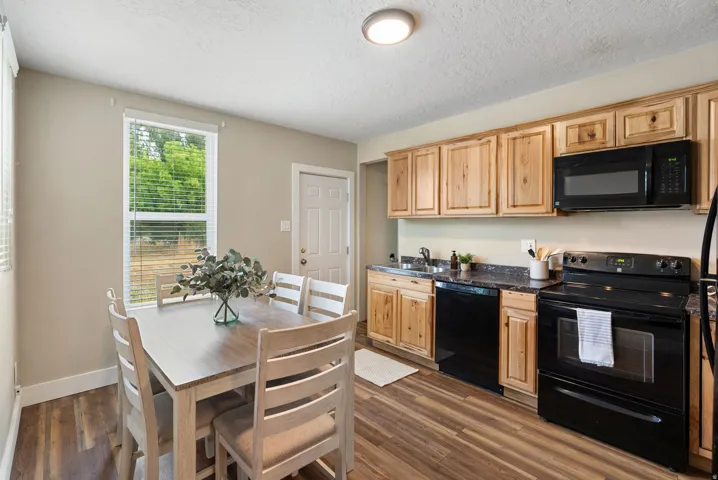 West unit. Integrated kitchen and dining space featuring wood-finish flooring, natural wood cabinetry, black appliances, dark countertops, and a window with blinds
