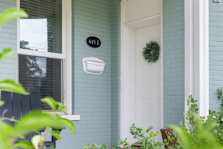 Entrance to the East unit. Front exterior showcasing light blue painted brick, a white paneled entry door, and a white window frame with blinds