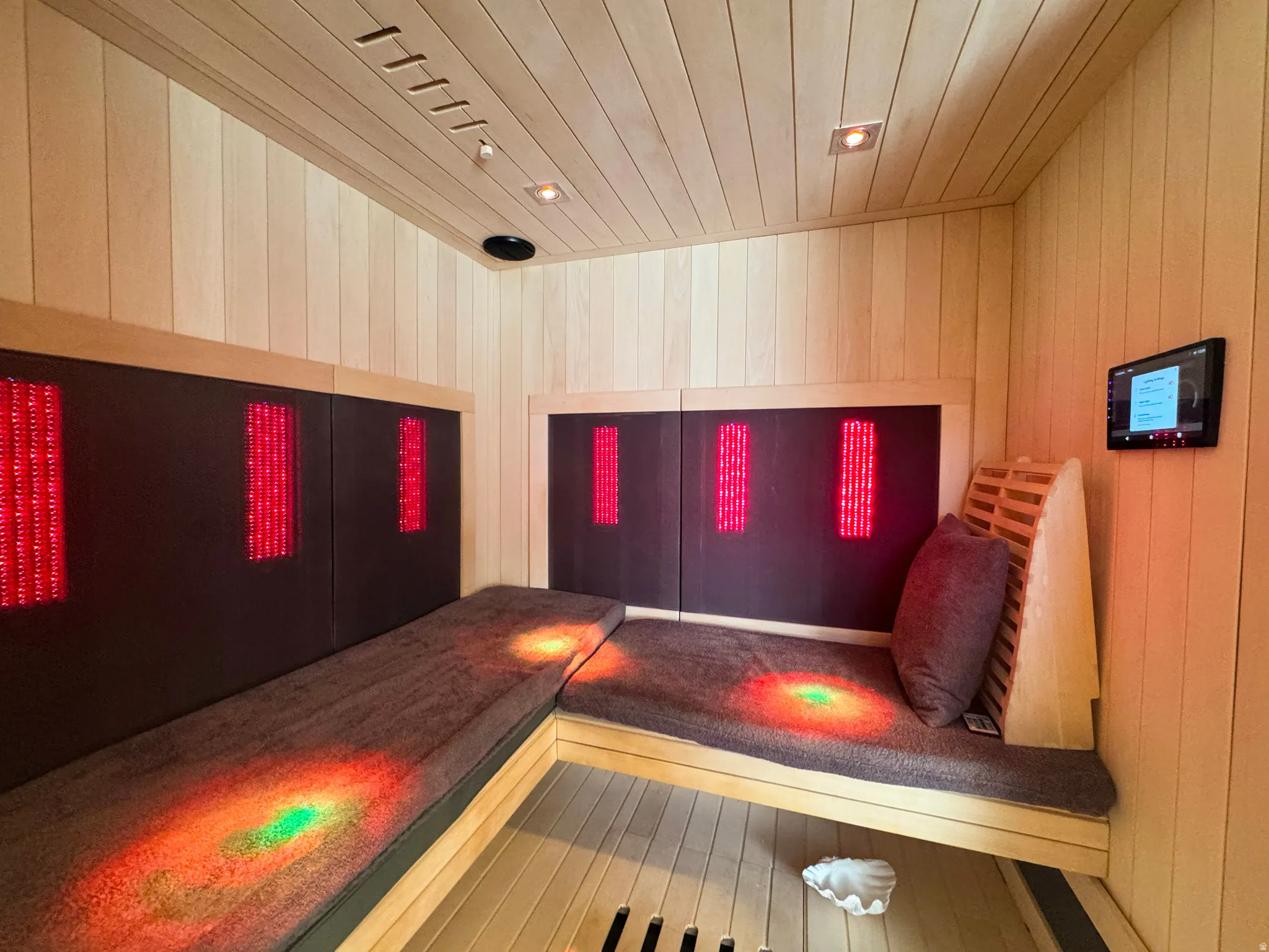 Custom-built sauna featuring light-toned wood-paneled walls and ceiling, built-in bench seating with plush cushions, integrated red light therapy panels, recessed ceiling lighting, and a wall-mounted control panel