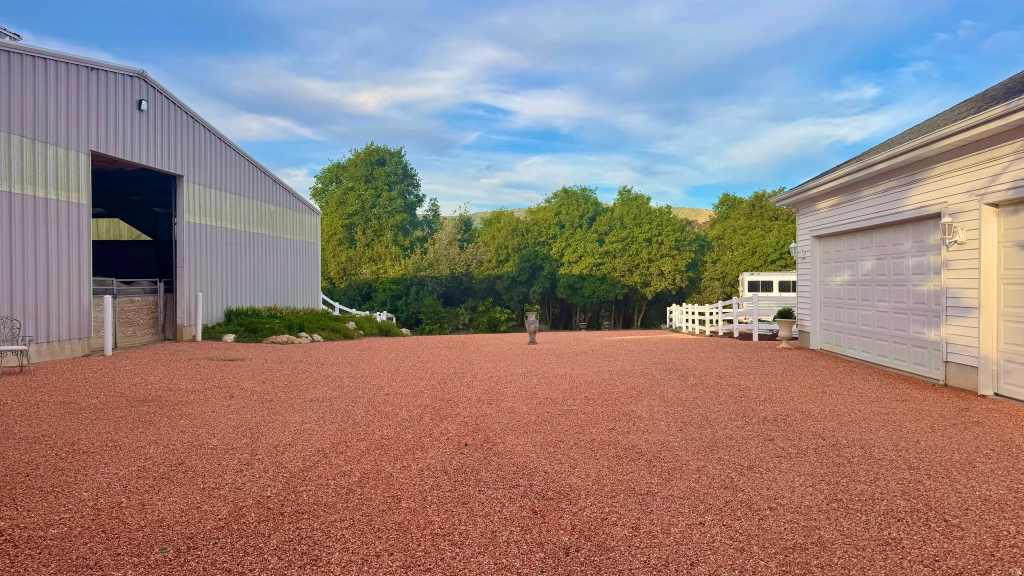 Expansive red gravel area with a large metal-sided outbuilding featuring an open entryway and a white-sided two-car garage with overhead doors
