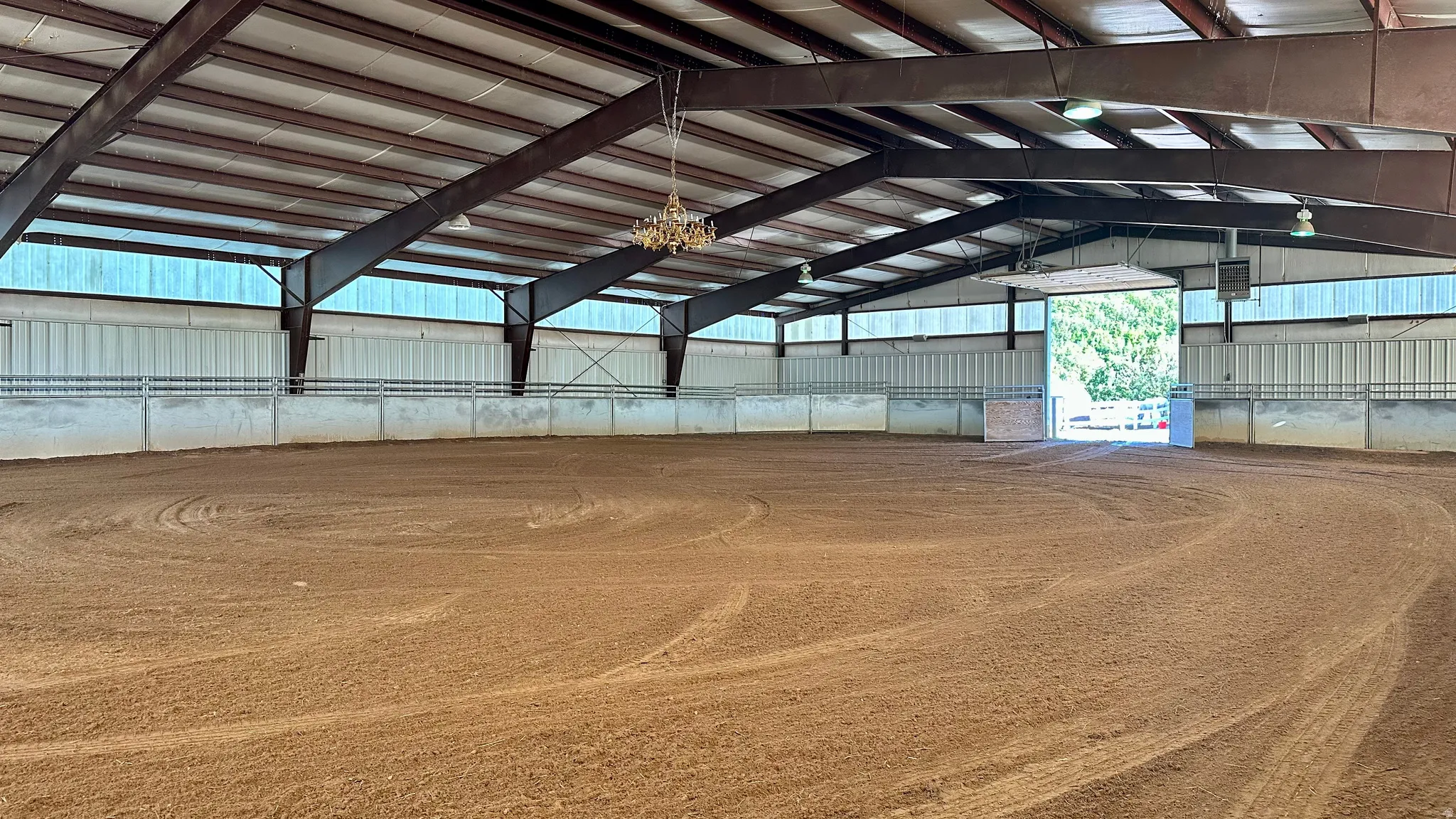 Expansive indoor arena featuring a sand surface, high ceilings with exposed steel beams, perimeter railing, and a large entry door