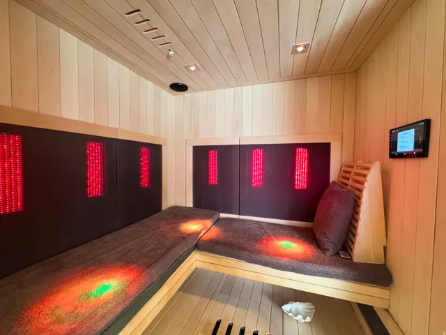 Custom-built sauna featuring light-toned wood-paneled walls and ceiling, built-in bench seating with plush cushions, integrated red light therapy panels, recessed ceiling lighting, and a wall-mounted control panel