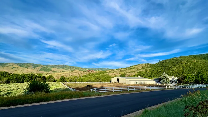 Expansive rural property featuring a large barn structure, a white-fenced perimeter, and a residential building with a prominent evergreen tree