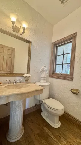 Pedestal sink with a stone finish, a wall-mounted light fixture, wood-finish flooring, a window with wood trim, and a white toilet