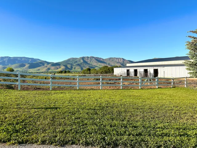 Expansive outdoor area featuring a white perimeter fence, a large metal-sided outbuilding with multiple stalls, and distant mountain scenery