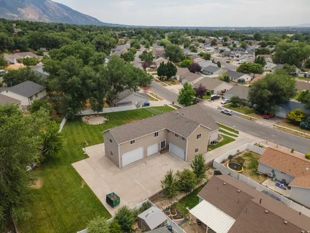 Expansive concrete driveway and two-story property featuring two garage doors, a large green lawn, mature trees, and a mountain backdrop