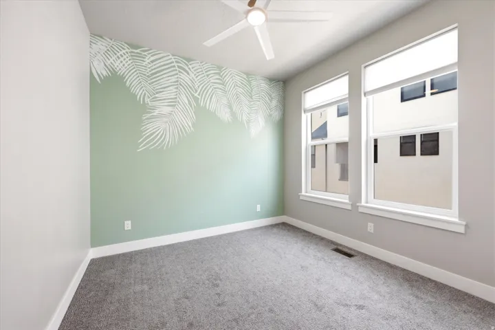 Room featuring vaulted ceilings, a ceiling fan, two large windows with roller shades, a decorative accent wall, and grey carpeting