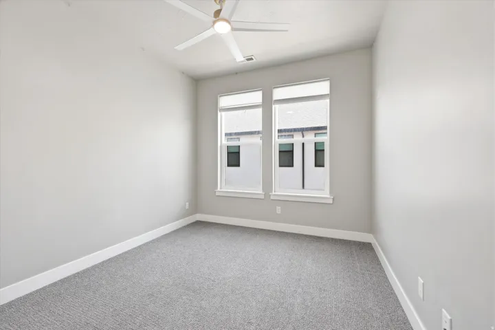 Bright interior room featuring a ceiling fan, two tall windows with roller blinds, light gray carpet, and white baseboards