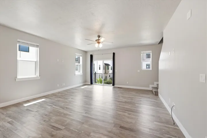 Spacious living area featuring light wood-finish flooring, multiple windows, a ceiling fan with integrated lighting, a sliding glass door opening to an exterior balcony with black railings, and a carpeted staircase