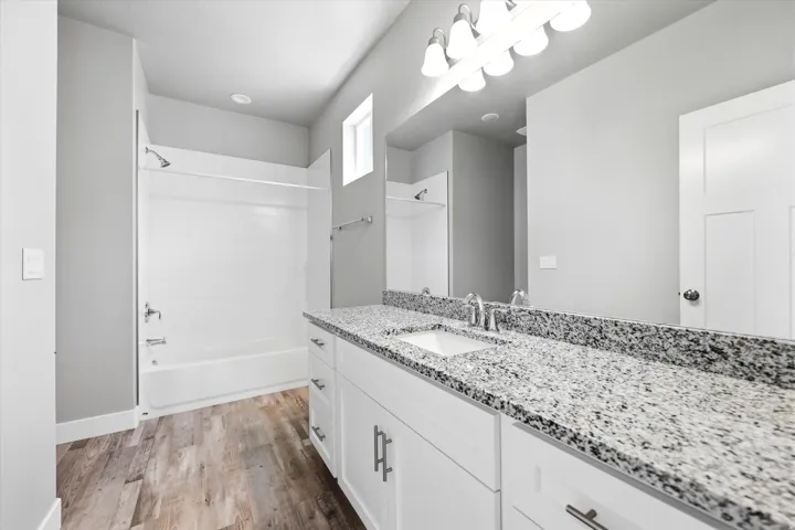 Bathroom featuring a white bathtub/shower combination, white vanity with dual sinks, granite countertop, wood-finish flooring, and a rectangular transom window