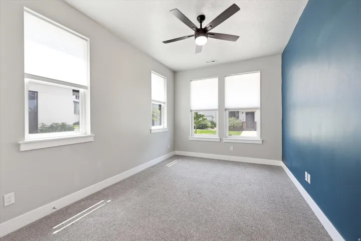 Bright interior space featuring a ceiling fan with integrated lighting, multiple windows with white blinds, a contrasting accent wall, and gray carpeting