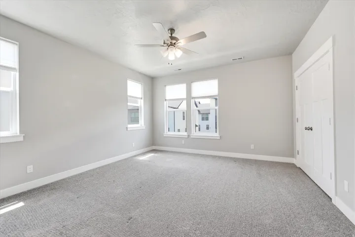 Spacious room featuring light gray carpeting, multiple windows with roller shades, a ceiling fan with integrated lighting, and a white interior door with dark hardware