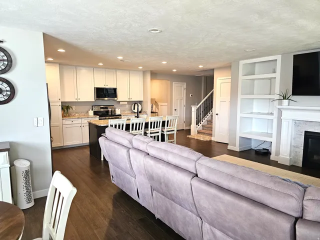 Open-concept living area featuring wood-finish flooring, a kitchen with white cabinetry and a center island, a fireplace with a tile surround, built-in shelving, and a staircase with a dark wood handrail
