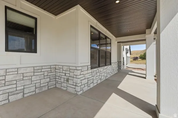 Covered concrete patio featuring stone veneer siding, white stucco, multiple windows with dark framing, and a dark wood-finish ceiling with recessed lighting