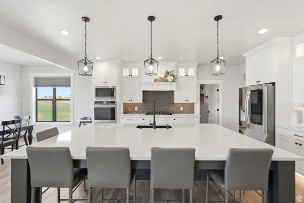 Spacious kitchen featuring a large central island with seating, white shaker cabinetry, stainless steel appliances, dark tile backsplash, and recessed and pendant lighting
