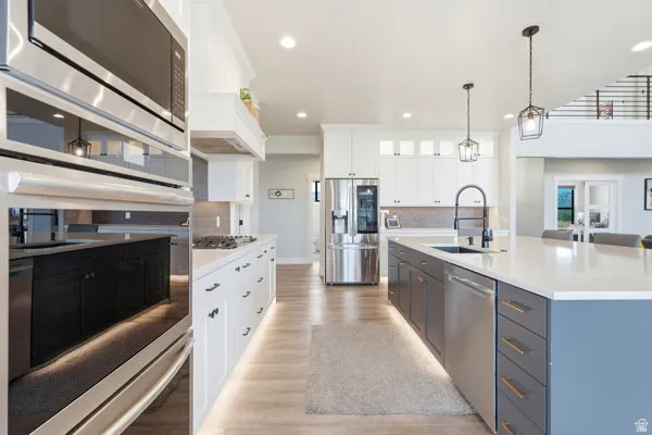Contemporary kitchen featuring a large central island with a sink and pendant lighting, stainless steel appliances including a refrigerator and wall oven, white cabinetry, and wood-finish flooring