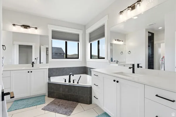 Spacious bathroom featuring dual vanities with white cabinetry and light-toned countertops, a corner soaking tub with dark tile surround, and a large mirror extending across the wall